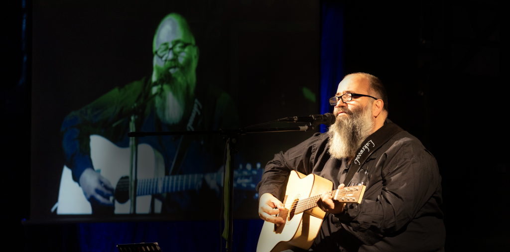 Micha Krämer beim Gitarre spielen und Singen auf einer Bühne mit einem Beamerbild im hintergrund auf dem er auch zu sehen ist.